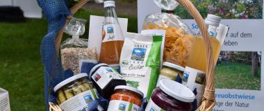 Ein Präsentkorb mit regionalen Lebensmitteln von „Unser Land“ steht im Freien. Im Korb befinden sich unter anderem Saft, Nudeln, Zucker, Haferflocken sowie Gläser mit eingelegtem Gemüse und Konfitüre. Im Hintergrund ist ein Informationsplakat zu regionalen Projekten zu sehen.